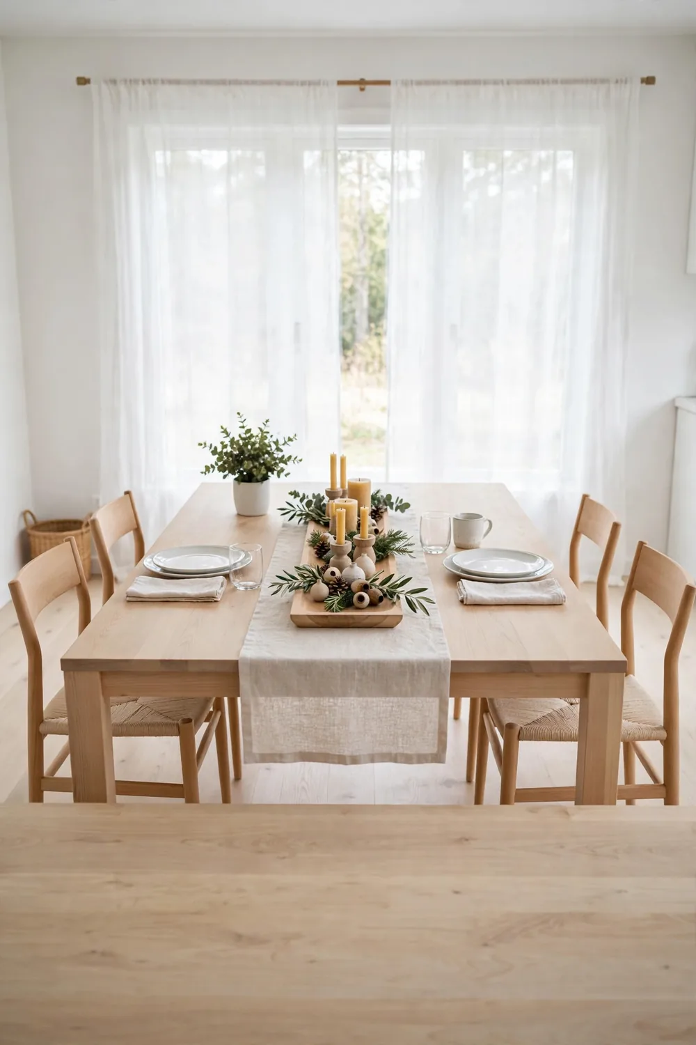 Long wooden tray holding candles, greenery and ornaments as a tidy centerpiece on a linen runner in the middle of a dining table in a calm dining room.