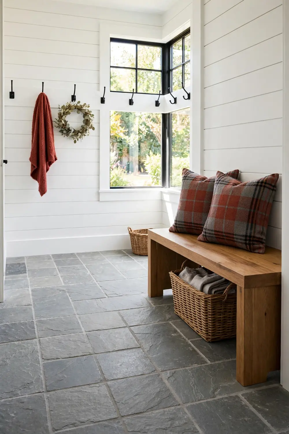 Wooden entry bench with tartan throw pillows and a woven basket beneath it in a bright mudroom style hall with shiplap walls and tile floors.